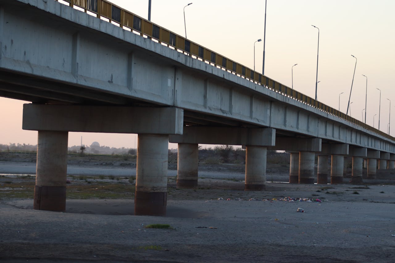 A concrete bridge spans across a dry riverbed at dusk, under a pastel sky.